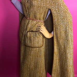 May include: A long, brown and white tweed cape with a brown leather belt and a brown pocket. The cape has a high collar and a long, flowing design.