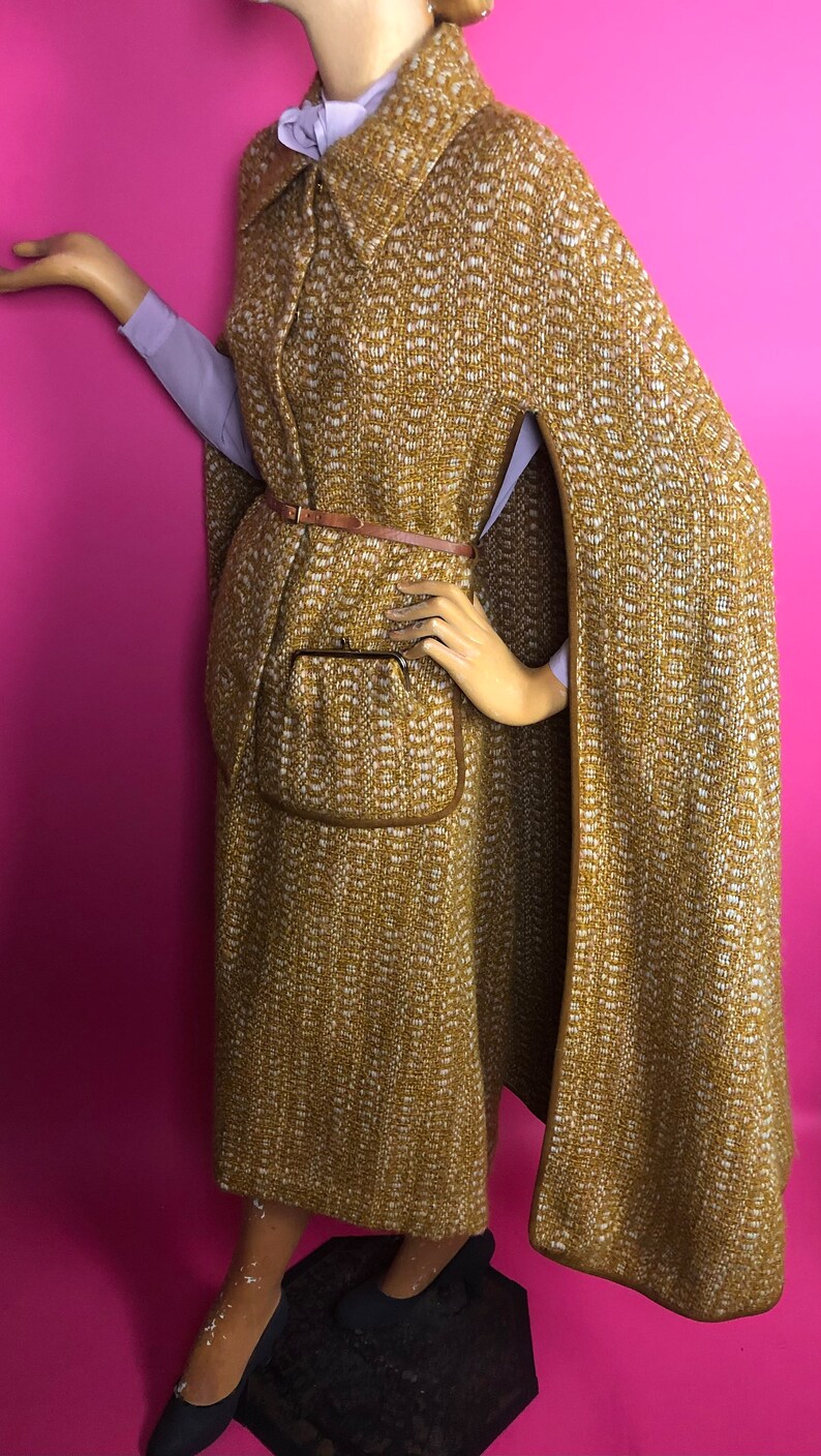 May include: A long, brown and white tweed cape with a brown leather belt and a brown pocket. The cape has a high collar and a long, flowing design.