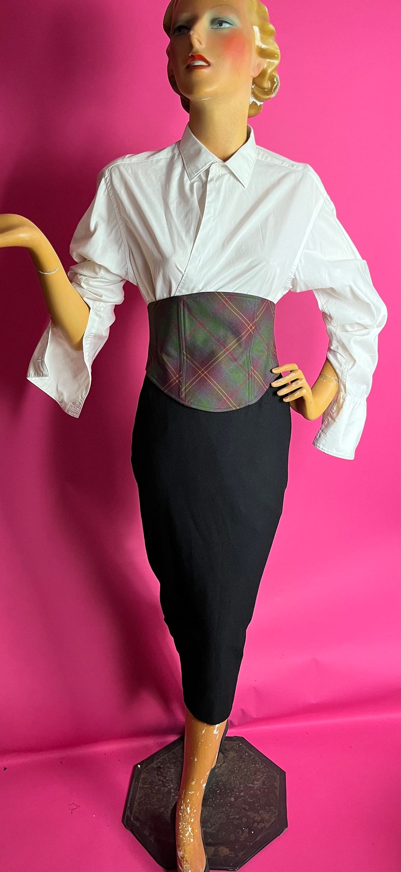 May include: A white button-down shirt with a plaid corset belt and a black pencil skirt. The mannequin is standing on a black octagonal base.