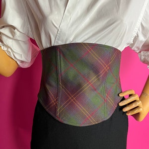 May include: A plaid corset with a green, brown, and red pattern. The corset is made of a soft, woven fabric and has a fitted silhouette.