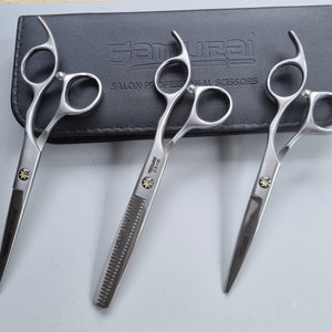 Puede incluir: Tres tijeras profesionales de peluquería Samurai plateadas con mangos negros y un estuche negro. Las tijeras incluyen dos tijeras estándar y unas tijeras de entresacar. El texto "Samurai" y "Salon Professional Scissors" es visible en el estuche.