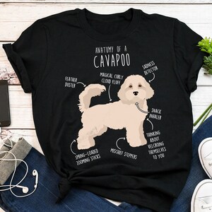 Puede incluir: Una camiseta negra con una ilustración blanca de un perro Cavapoo. La ilustración está etiquetada con características anatómicas humorísticas, como "nube de pelusa rizada mágica", "pasos traviesos" e "inhalador de bocadillos".