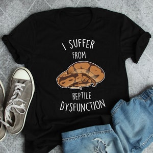 Könnte beinhalten: Schwarzes T-Shirt mit einem weißen Aufdruck, der "Ich leide unter Reptiliendysfunktion" sagt. Eine braune und beige Schlange ist auf dem T-Shirt zusammengerollt.