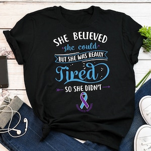 May include: Black t-shirt with a blue and purple ribbon and the text "SHE BELIEVED she could BUT SHE WAS REALLY Tired SO SHE DIDN'T" with arrows pointing to the left and right of the text. The ribbon has the words "RHEUMATOID ARTHRITIS" written on it.