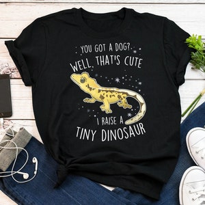 Op de afbeelding: Zwart T-shirt met een witte afbeelding van een gekko-hagedis en de tekst "You got a dog? Well, that's cute. I raise a tiny dinosaur."