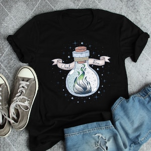 以下が含まれることがあります： 緑と白の炎が入ったポーションボトルのグラフィックが特徴の黒いTシャツ。ボトルにはコルク栓があり、バナーには「Soul Fire」と書かれています。グレーのスニーカーとブルージーンズも写っています。