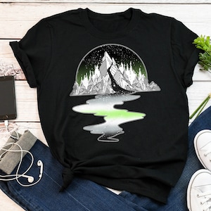 May include: Black t-shirt with a graphic of a mountain range reflected in a body of water. The water is green, white, and gray, representing the colors of the ace flag.