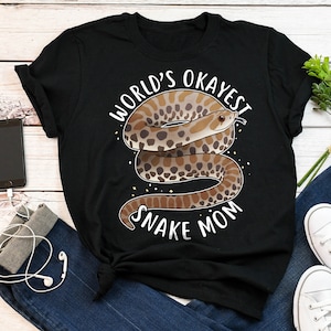 Puede incluir: Camiseta negra con una ilustración de serpiente marrón y blanca y el texto "World's Okayest Snake Mom".