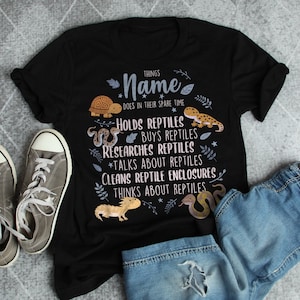 Op de afbeelding: Zwart T-shirt met een witte afbeelding waarop staat "Things Name Does In Their Spare Time" en activiteiten worden vermeld die verband houden met reptielen. De afbeelding bevat illustraties van een schildpad, een slang en een hagedis.