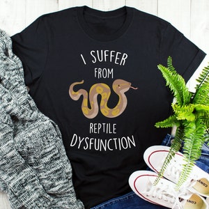 May include: Black t-shirt with the text "I Suffer From Reptile Dysfunction" and a cartoon snake illustration. The snake is brown and yellow. The shirt is paired with white sneakers and a gray sweater.