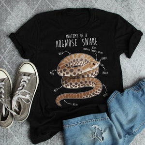 Peut inclure: Un t-shirt noir avec une illustration blanche d'un serpent à nez de cochon. L'illustration comprend le texte "Anatomie d'un serpent à nez de cochon" et des étiquettes pour le cou, le museau et le pbbbt du serpent.
