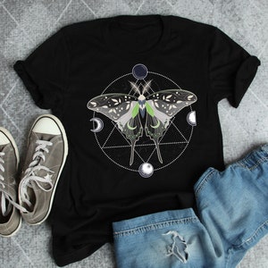 May include: A black t-shirt with a graphic of a green and white butterfly inside a circle with moons and stars. The butterfly has a geometric pattern on its wings.