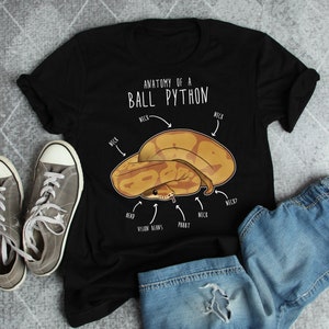 Op de afbeelding: Een zwart T-shirt met een witte afbeelding van een ball python met de tekst "Anatomie van een ball python" en labels voor de kop, zichtbonen, pbbbt, nek en nek?