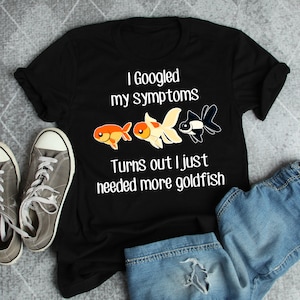 May include: Black t-shirt with white text that reads "I Googled my symptoms Turns out I just needed more goldfish". Three goldfish are illustrated on the shirt, two orange and one black and white.