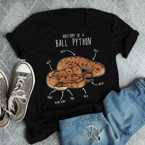May include: Black t-shirt featuring an illustrated ball python with labeled anatomy. The text "ANATOMY OF A BALL PYTHON" is above the snake. Gray canvas sneakers and denim shorts are also in the image.