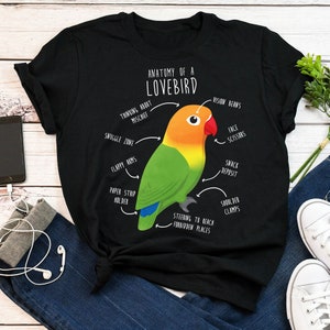 Pode incluir: Camiseta preta com uma ilustração humorística de um agapornis com características anatômicas etiquetadas, incluindo "Thinking About Mischief", "Vision Beans", "Face Scissors", "Snack Deposit", "Shoulder Clamps", "Steering to Reach Forbidden Places", "Smuggle Zone", "Flappy Arms", e "Paper Strip Holder".