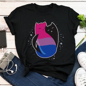 May include: Black t-shirt with a graphic of two cats cuddling. The cat on the left is pink, blue, and purple, and the cat on the right is black. The graphic is surrounded by white stars.