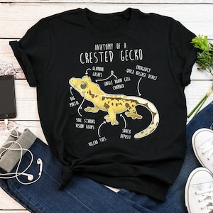 Peut inclure: T-shirt noir avec une illustration blanche d'un gecko à crête avec des détails anatomiques étiquetés. Le texte indique "Anatomie d'un gecko à crête" avec des étiquettes telles que "Cils glamour", "Portail des insectes", "Vision des haricots fixant l'âme", "Chambre à cellule cérébrale unique", "Dispositif de libération rapide", "Dépôt de collations" et "Doigts velcro".