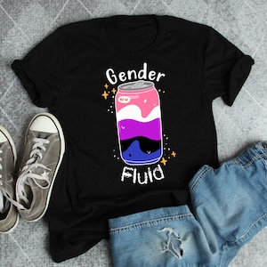 Puede incluir: Camiseta negra con un gráfico de una lata de refresco etiquetada como "Gender Fluid". La lata está decorada con los colores de la bandera del orgullo genderfluid: rosa, blanco, morado, azul y negro.