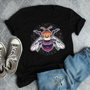 Puede incluir: Camiseta negra con un gráfico de una abeja con una luna creciente en la cabeza. La abeja está rodeada de un círculo de estrellas. La abeja está coloreada en tonos de rosa, morado y naranja.