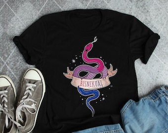 Può includere: Una maglietta nera con una grafica di due serpenti intrecciati, uno rosa e viola e l'altro blu. I serpenti sono avvolti attorno a uno stendardo che recita "BISNEKUAL".