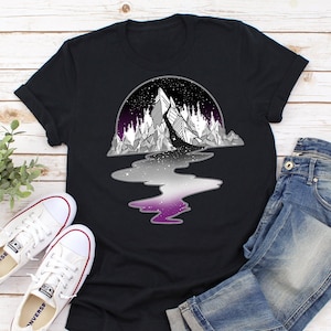 Könnte beinhalten: Schwarzes T-Shirt mit einem Grafikdruck einer Bergkette, die sich in einem Fluss mit einem Farbverlauf in Lila, Weiß und Schwarz spiegelt. Der Fluss ist mit Sternen gefüllt.