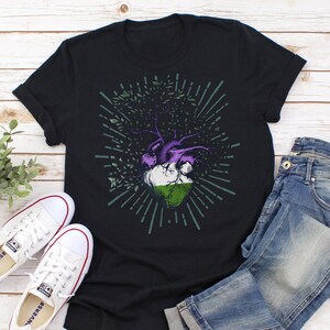 May include: A black t-shirt with a graphic of a heart with a tree growing out of it. The heart is colored in the colors of the genderqueer pride flag: purple, white, and green. The tree has green leaves and a purple trunk. The graphic is surrounded by a circle of green rays.