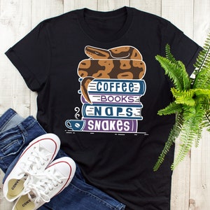 May include: Black t-shirt with a graphic of a snake curled up on a stack of books. The books have the words "Coffee", "Books", "Naps", and "Snakes" written on them. There is a small cup of coffee next to the books.