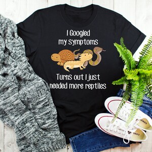 May include: Black t-shirt with a white graphic of a turtle, a bearded dragon, and a snake. The text on the shirt reads "I Googled my symptoms Turns out I just needed more reptiles."