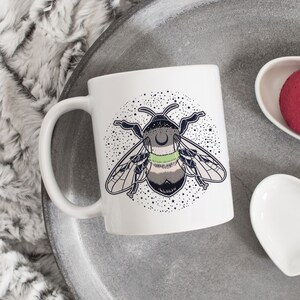 May include: White ceramic mug featuring a black and green bee illustration. The bee has a crescent moon on its head and is surrounded by a circle of stars. The mug is on a grey tray with two white heart-shaped dishes.