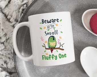 Op de afbeelding: Een witte keramische mok met een illustratie van een groene en blauwe vogel en de tekst "Beware of the Small Fluffy One".