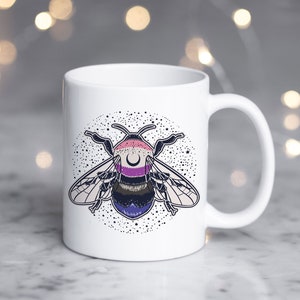 Puede incluir: Taza de cerámica blanca con una ilustración en blanco y negro de una abeja con una franja rosa, morada y blanca en el abdomen. La abeja está rodeada por un círculo de estrellas.