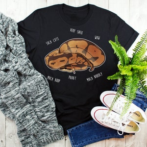 Könnte beinhalten: Schwarzes T-Shirt mit einer Cartoon-Illustration einer braunen und braunen Schlange. Die Schlange ist zusammengerollt und hat einen süßen Ausdruck. Der Text um die Schlange lautet "Very Snek", "Such Cute", "Wow", "Much Boop", "Pbbbt" und "Much Woodly".