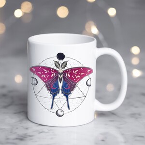 May include: White ceramic mug with a pink and blue butterfly design inside a circle with moons and a geometric pattern.