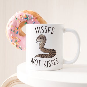 May include: White ceramic coffee mug with a brown snake illustration and the text "Hisses not Kisses". A pink frosted donut with colourful sprinkles is behind the mug.