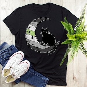 May include: Black t-shirt with a black cat sitting on a crescent moon. The moon is decorated with grey, white, and green stripes.