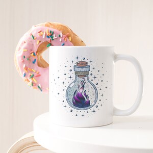 Puede incluir: Taza de cerámica blanca con un gráfico de una llama negra y morada dentro de una botella de vidrio. La botella tiene un tapón de corcho y está rodeada de estrellas blancas. La taza está sobre una superficie blanca.
