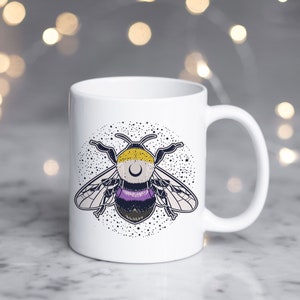 May include: White ceramic mug featuring a stylized bee design with yellow, purple, and black accents. The bee is set against a backdrop of small white stars. The mug has a curved handle and a glossy finish.