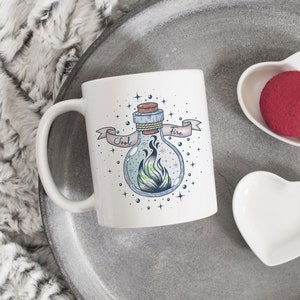 Puede incluir: Taza de cerámica blanca con un gráfico de una botella de vidrio con un tapón de corcho y una llama en su interior. La botella tiene las palabras "Soul Fire" escritas en una cinta alrededor de ella. La taza está sobre una bandeja gris con dos platos blancos en forma de corazón.