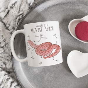 May include: White ceramic mug with a cartoon illustration of a hognose snake with the text "Anatomy of a Hognose Snake" and "Pranatic Thoughts Inside".