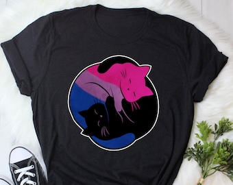 Può includere: Maglietta nera con un design yin yang con due gatti. I gatti sono di colore rosa, nero e blu, a rappresentare la bandiera del pride bisessuale.