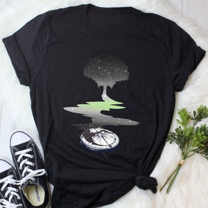 Könnte beinhalten: Schwarzes T-Shirt mit einem Motiv eines Baumes mit einem Sternenhimmel in seinen Ästen, der sich in einer Wasserpfütze mit einem Kompass am Boden spiegelt.