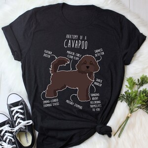 Könnte beinhalten: Ein schwarzes T-Shirt mit einer humorvollen Illustration eines Cavapoo-Hundes. Die Illustration ist mit lustigen anatomischen Merkmalen wie "magischer lockiger Wolkenflaum" und "Snack-Inhalator" beschriftet.