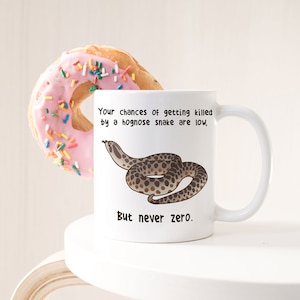 May include: White ceramic coffee mug featuring a brown and tan hognose snake illustration. The mug reads "Your chances of getting killed by a hognose snake are low, But never zero."