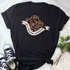 Op de afbeelding: Zwart T-shirt met een afbeelding van een bruine en beige slang die om een wit lint gewikkeld is met de tekst "I Love Danger Noodles".