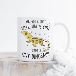 May include: White ceramic mug featuring a cartoon gecko and the text "You got a dog? Well, that's cute. I raise a tiny dinosaur."