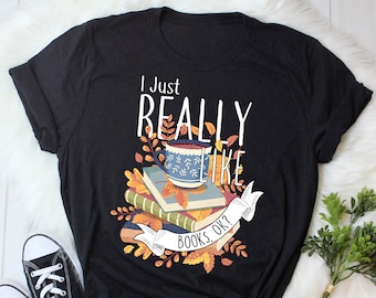 Op de afbeelding: Zwart T-shirt met een afbeelding van een stapel boeken, een kopje koffie en herfstbladeren. De tekst op het shirt luidt "I just really like books, ok?"