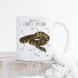 May include: White ceramic mug featuring a cartoon illustration of a brown and tan snake. The mug is labelled "Anatomy of a Carpet Python" and includes labels for the snake's neck, head, and vision beans.