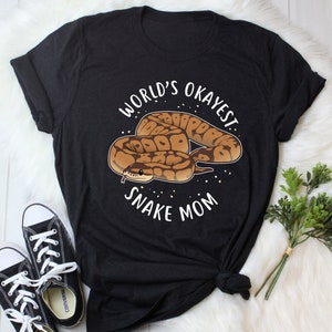 Pode incluir: Camiseta preta com um gráfico branco de uma cobra e o texto "World's Okayest Snake Mom".