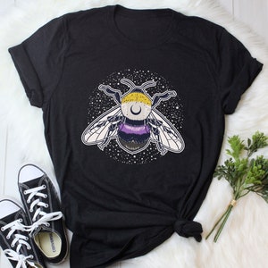 Peut inclure: T-shirt noir avec un motif d'abeille avec des rayures jaunes, violettes et noires. L'abeille est entourée d'étoiles blanches sur un fond noir. L'abeille a un croissant de lune sur la tête.
